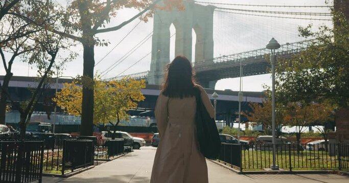 Happy and stylish brunette joyfully walks toward the iconic Brooklyn Bridge in an autumn sunny New York City landscape