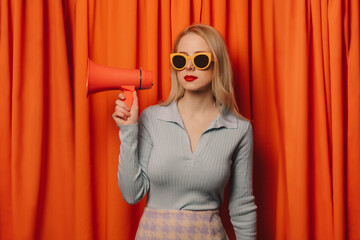 Woman wearing sunglasses holding megaphone in front of orange curtain
