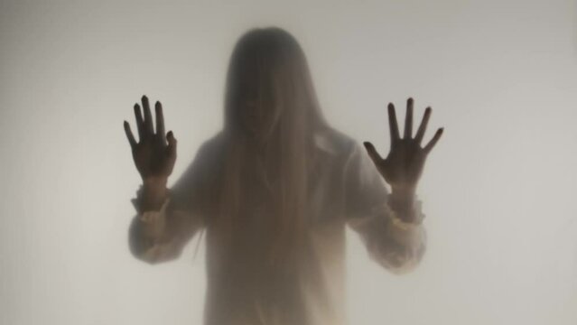 Silhouette of panicked woman in fog behind frosted glass or curtain. The woman bangs her head against a transparent, invisible barrier. Concept of the afterlife and the otherworld, ghosts and spirits
