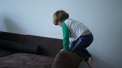 One small boy climbs on sofa edge playing at home. Child mounting on top of couch