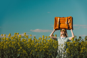 Happy blond woman with suitcase on head in rapeseed field