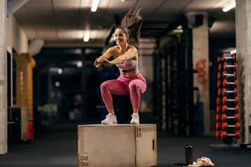 A happy sportswoman is jumping on a jump box in a gym with heart rate belt.