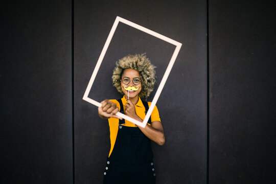 Woman holding fake mustache and frame in front of black wall