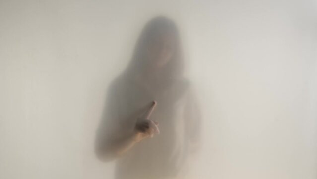 Silhouette of ghost woman in fog behind frosted curtain or glass close up. The woman draws invisible patterns on the glass with her index finger. The concept of the afterlife, the otherworld, ghosts