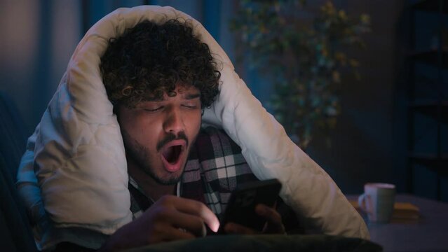 Arabian exhausted yawning guy using mobile phone insomnia night Indian man fatigue yawn tired sleepy late under blanket male reading social media app bad habit scrolling smartphone sleepless addiction