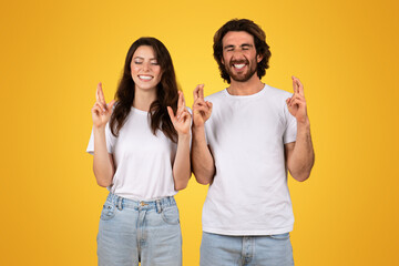 Optimistic couple with closed eyes crossing their fingers for good luck, both wearing white t-shirts