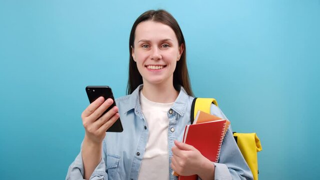 Portrait of excited woman student in shirt with backpack hold notebooks using mobile cell phone typing sms message, isolated over blue background. Education in high school university college concept