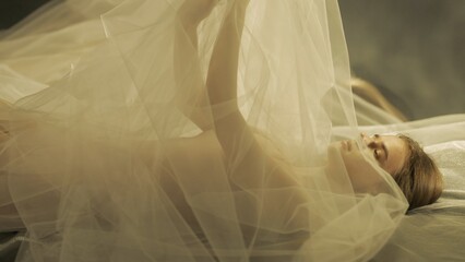 Side view of a young woman on a bed, under a transparent air cloth, close up. A woman illuminated by warm light raised her arms, forming a tulle like canopy. - obrazy, fototapety, plakaty