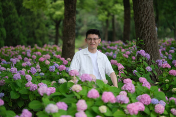 handsome Asian young man in glasses at colorful hydrangea macrophylla flowers, front portrait