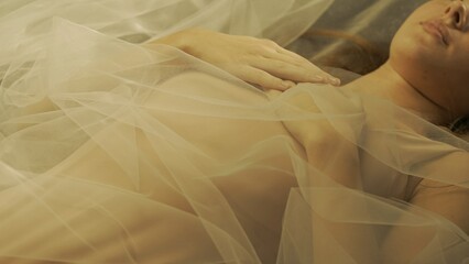 Hands of woman with nude manicure, folded on woman's chest, close up. Woman lying on her back covered with white tulle, illuminated by warm rays of light.