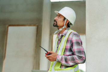 Male construction engineer uses walkie-talkie communicate with other builder in same construction area, communicate with words gestures tell builder needs in order complete planned house plan.