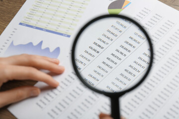 Woman looking at accounting document through magnifying glass at table, closeup