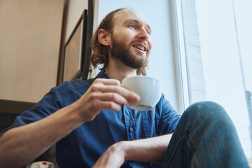 View from below. Young man sitting and looking to window and drinking hot coffee or tea. Morning routine. Concept of beauty and medicine, health care, lifestyle. Perfect smile.
