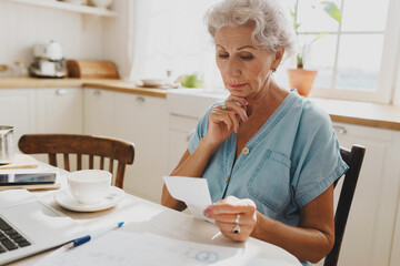 Puzzled worried serious elderly lady accountant with gray hair working from home, examining pay...