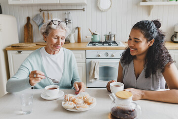 Indoor portrait of two people of diverse age and ethnicity sitting at kitchen table, african american young woman drinking tea with elderly caucasian female treating her with sweets - obrazy, fototapety, plakaty