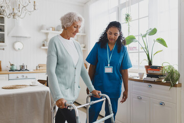 Side view of black african american nurse volunteer teaching elderly woman with gray hair in cardigan to use walker, supporting her and giving advice and instructions, standing in her kitchen