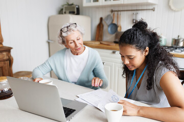 Black girl volunteer or social worker helping elderly lonely woman to deal with documents, agreement or invoice, reading and examining paper doc sitting at kitchen table together in front of laptop