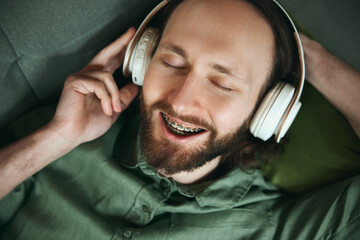 Vie from above. Joyful young man in braces lying on couch and listening music in headphones and singing. Perfect smile and delight customer. Concept of beuty and healthcare, medicine, lifestyle.