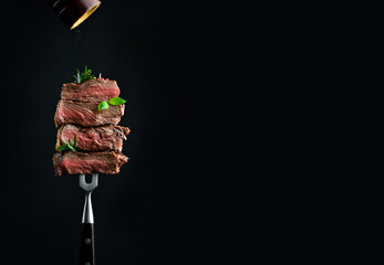 Beef steak on a fork with a pepper mill, frozen pepper particles in the photo. Grilled steak. On a black background. © Yaruniv-Studio