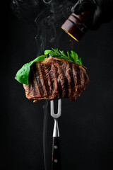 Beef steak on a fork with a pepper mill, frozen pepper particles in the photo. Grilled steak. On a black background. - obrazy, fototapety, plakaty