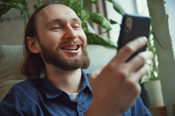 Delightful bearded young man with braces sitting with phone in comfy armchair and scrolling social...