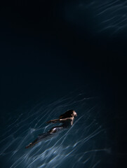 Underwater shoot of beautiful girl in white dress swimming in water through sunbeams.