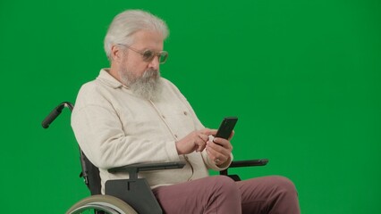 Portrait of disabled man isolated on chroma key green screen close up. Senior man sitting in wheelchair holding smartphone watching video.