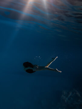 Underwater shoot of beautiful sportive girl in bodysuit swimming in water through sunbeams.