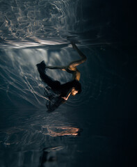 Underwater shoot of ballerina in black dress swimming and dancing in water through sunbeams.