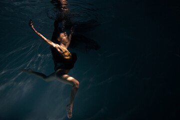 Underwater shoot of ballerina in black dress swimming and dancing in water through sunbeams.