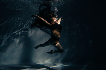 Underwater shoot of ballerina in black dress swimming and dancing in water through sunbeams.