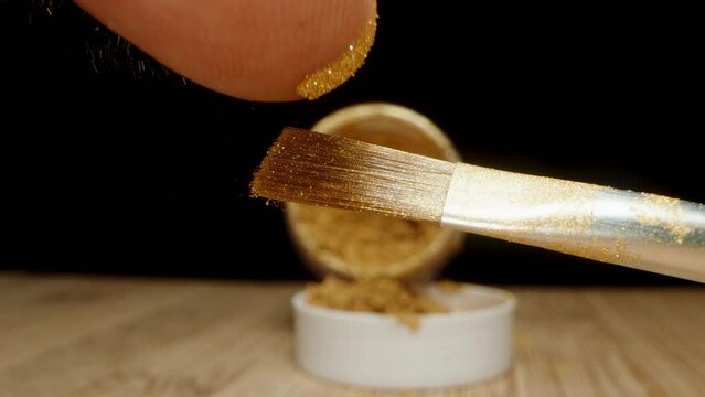 Close-up of a brush with golden particles of powder or dust on it, being flicked by a finger and scattering in the air, against a black background.