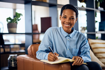 A portrait of a smiling black woman, sitting on the sofa in her office, writing some notes in her...