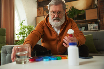 Senior man taking care after health with meds, vitamins. Man sitting at home, taking glass of water...