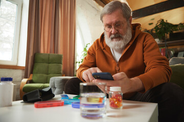 Focused elderly man with glasses using smartphone, checking medical prescriptions, contacting with doctor. Pill organizer and water glass on table. Concept of health and medical care, aging, medicine