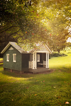 A charming small cottage with a green exterior and white trim sits peacefully under a tree in a sunlit garden. The lush grass and dappled sunlight create a serene and inviting atmosphere.