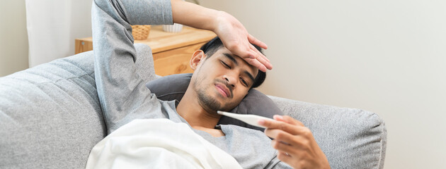 Seasonal cold sickness in people concept, Sick asian man looking at thermometer during measuring...