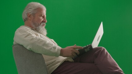 Portrait of aged bearded man on chroma key green screen background. Full shot of senior man sitting on a chair working on laptop.