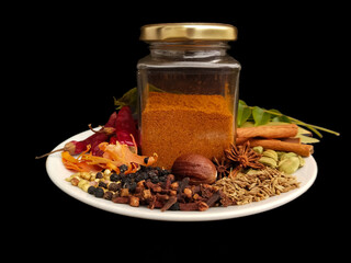 Garam masala powder in a glass bottle with ingredients on a plate, black background