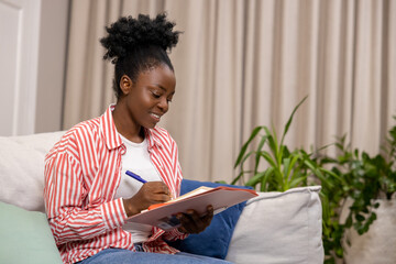 Woman writing her personal notes into her diary while sitting on sofa in living room.