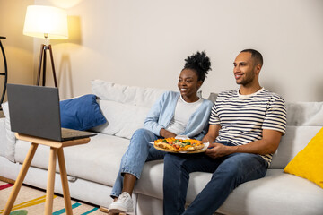 Male and female couple eating pizza while watching movie on laptop and sitting on couch