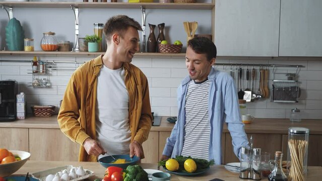 A cheerful male gay couple preparing breakfast together in the kitchen. Happy attractive young adult men joyfully spending time together cooking delicious dishes side by side. High quality 4k footage