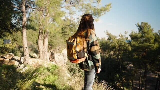 A young woman with a backpack in the forest, enjoying the view from a high cliff with the sea and pine trees in the background.