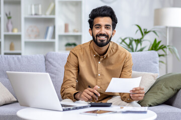 Close-up portrait of a young Indian man working remotely from home, sitting on a sofa, holding documents and using a calculator, smiling at the camera.