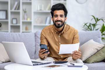 Portrait of a young Indian man sitting at home on the couch, holding a phone and documents, bills. Looking and smiling at the camera.