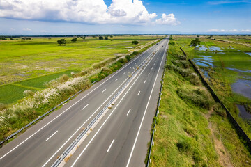 Fototapeta premium Scenic aerial shot of an empty highway cutting through lush green fields under a clear blue sky, emanating tranquility and ease. At Central Luzon Link Expressway.