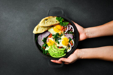 Female hands hold a pan with Shakshuka eggs with toast on a black concrete background. Traditional Jewish scrambled eggs.