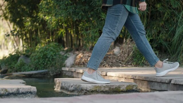 Female legs in jeans and sneakers stepping on stones across a small stream. Slow motion.