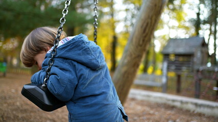 Contemplative Autumn Play - Thoughtful Child on Park Swing plays by himself while leaning on swing, twisting and turning amidst orange foliage
