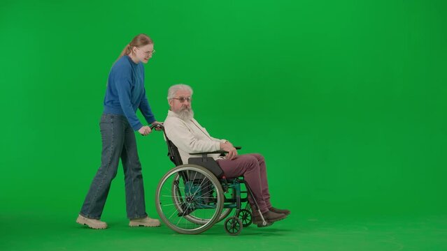 Portrait of aged disabled man on chroma key green screen. Girl granddaughter carries senior man on a wheelchair and talking.